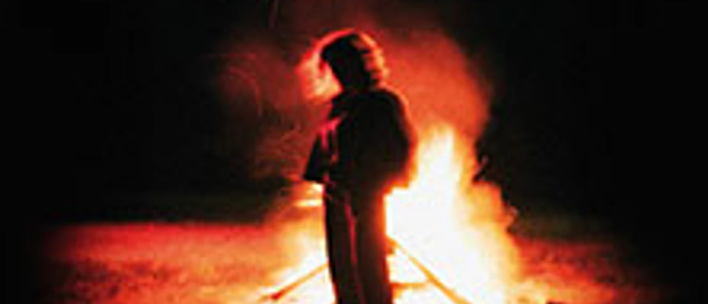 Silhouette vor großem Feuer in dunkler Landschaft, umgeben von rötlichem Lichtschein.
