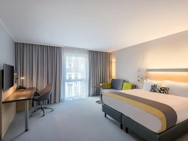 Modernes Hotelzimmer mit Doppelbett, Schreibtisch, TV, Sitzbereich und großem Fenster.