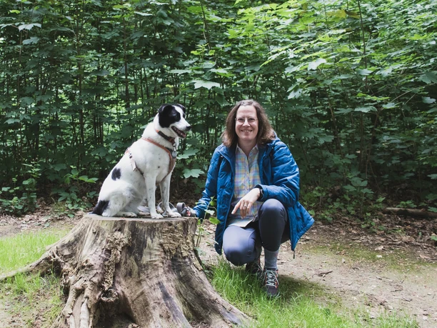 After-Work Tour auf der Entdeckerschleife Denkmalroute Hochdahl Lächelnde Wanderin mit Hund in grüner Waldlandschaft, der Hund auf einem Baumstamm sitzend.