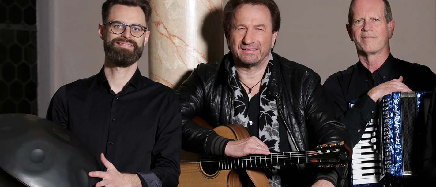 Clemens Bittlinger & Freunde Drei Musiker mit Gitarre, Handpan und Akkordeon in einer Kirche, bereit für ein Konzertauftritt.