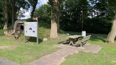Rastplatz-Apen-Nordloh-Plaggenhütte.jpg Holzhütte mit Infotafel neben Picknickbank und Tisch auf grünem Rastplatz unter hohen Bäumen.