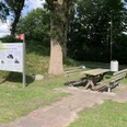 Rastplatz-Apen-Nordloh-Plaggenhütte.jpg Holzhütte mit Infotafel neben Picknickbank und Tisch auf grünem Rastplatz unter hohen Bäumen.