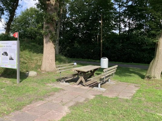 Rastplatz-Apen-Nordloh-Plaggenhütte.jpg Holzhütte mit Infotafel neben Picknickbank und Tisch auf grünem Rastplatz unter hohen Bäumen.