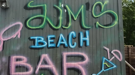 2024_Jims-Beachbar (5)_HD_16zu9.jpg Bunte Graffiti-Schrift mit dem Namen JIM’s Beach Bar auf grauer Holzfassade mit Cocktailmotiv.