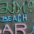 2024_Jims-Beachbar (5)_HD_16zu9.jpg Bunte Graffiti-Schrift mit dem Namen JIM’s Beach Bar auf grauer Holzfassade mit Cocktailmotiv.