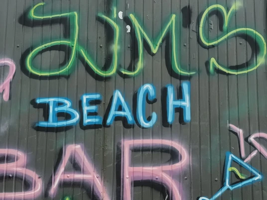 2024_Jims-Beachbar (5)_HD_16zu9.jpg Bunte Graffiti-Schrift mit dem Namen JIM’s Beach Bar auf grauer Holzfassade mit Cocktailmotiv.