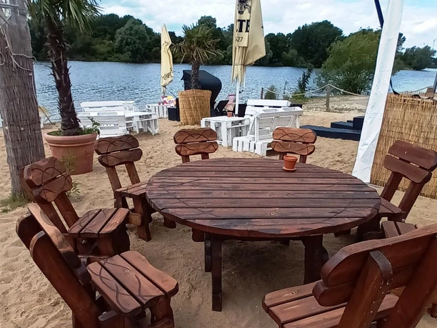 2024_Jims-Beachbar (6)_HD_16zu9.jpg Holztisch mit Stühlen im Sand, Palmen und Blick auf See mit weißen Sitzgruppen und Sonnenschirmen.