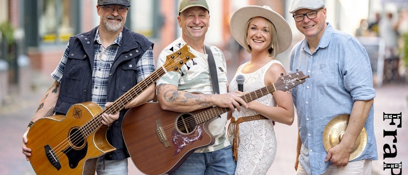 frapee.jpeg Gruppe von vier Personen auf gepflasterter Straße, zwei mit Akustikgitarren, eine mit Mikrofon, eine mit Becken, im Hintergrund unscharfe Gebäude