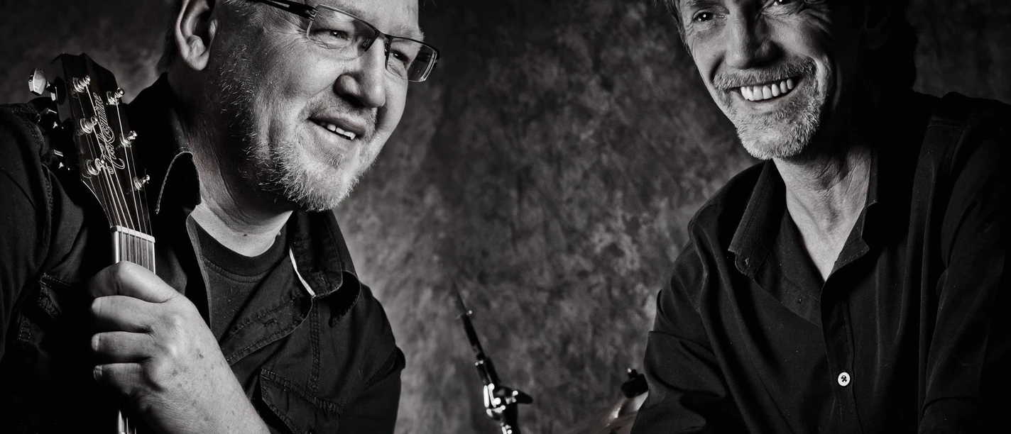 Querbeat2.jpeg Zwei Männer in schwarzer Kleidung, einer hält eine Gitarre, der andere lehnt sich mit verschränkten Armen auf ein Schlagzeug.