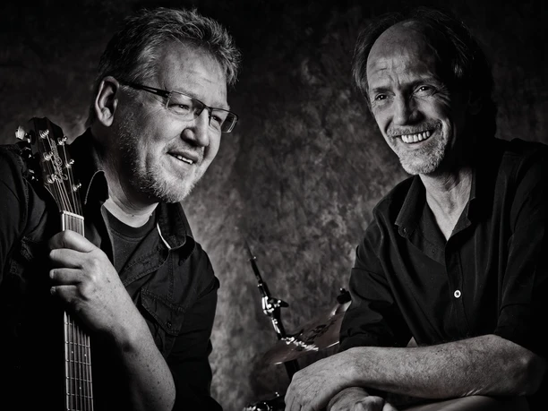 Querbeat2.jpeg Zwei Männer in schwarzer Kleidung, einer hält eine Gitarre, der andere lehnt sich mit verschränkten Armen auf ein Schlagzeug.