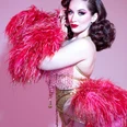 Medianoche Burlesque artist in a golden outfit with a pink feather boa against a pink background.