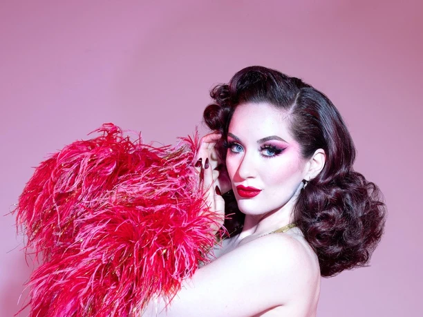 Medianoche Burlesque artist in a golden outfit with a pink feather boa against a pink background.