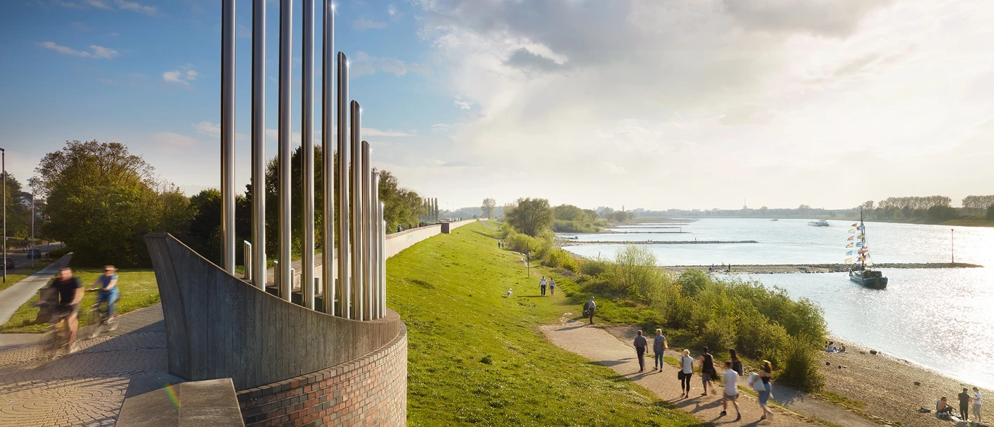 Feierabend am Rhein – Wandern & Genießen in Monheim Spaziergänger:innen und Radfahrer:innen entlang des Rheins in Monheim, mit Skulptur und Boot.