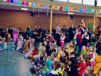 Kinderkarneval 2025.jpg Verkleidete Kinder sitzen in einer Aula, bunte Ballons darüber, ein Zauberer vorne