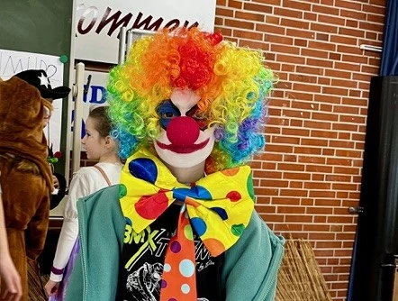 Clown beim Kinderkarneval in Apen 2025_Jörg Brandes.jpg Bunt verkleideter Clown mit Regenbogenperücke, roter Nase und großer Schleife in der Turnhalle.