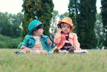 Zwei Kinder in farblich passenden Jäckchen und Hüten sitzen auf einer Wiese, das rechte Kind hat ein Telefon am Ohr.