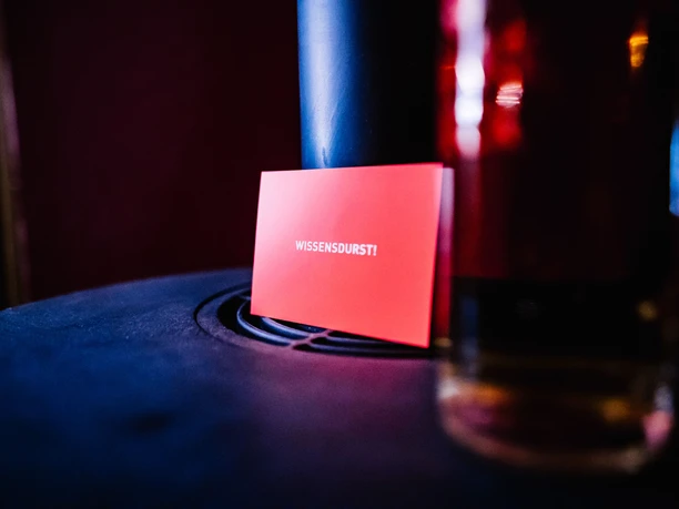 SGP_Fehrfeld_Ruth_Muentinga_Timm_Kroeger_17MAR22-013_2068.jpg Rote Karte mit Aufschrift WISSENSDURST! liegt auf dunklem Tisch, Glas unscharf im Vordergrund.