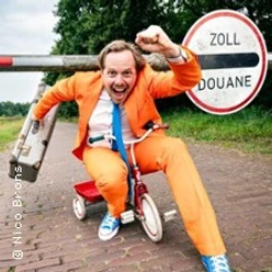 Mann in orangefarbenem Anzug fährt auf kleinem Fahrrad vor einem Zollschild über Kopfsteinweg.