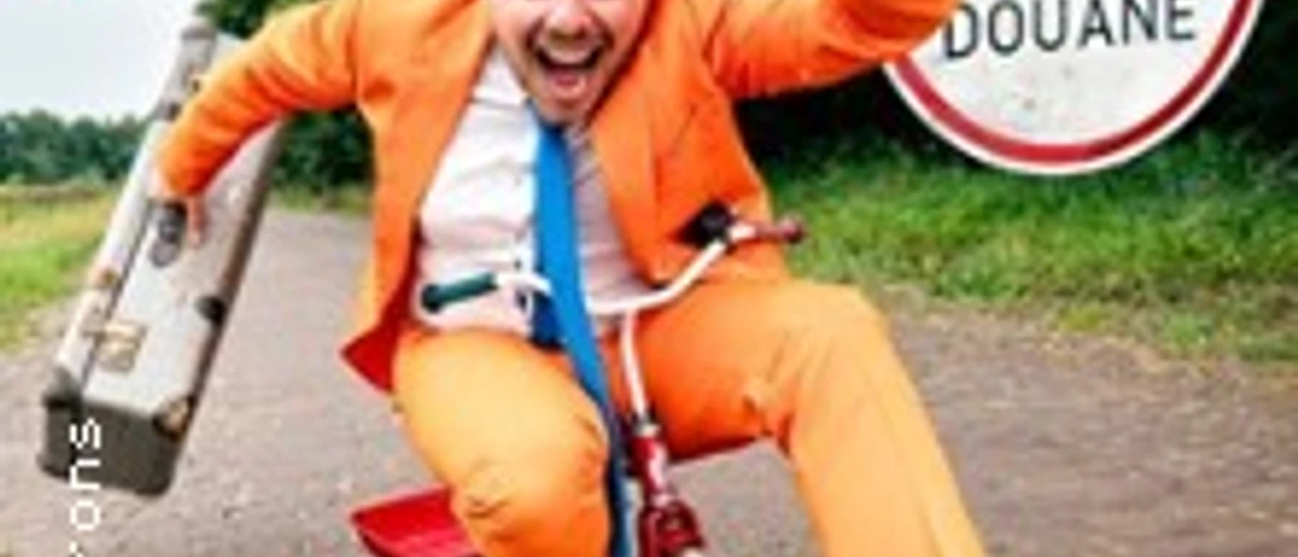 Mann in orangefarbenem Anzug fährt auf kleinem Fahrrad vor einem Zollschild über Kopfsteinweg.