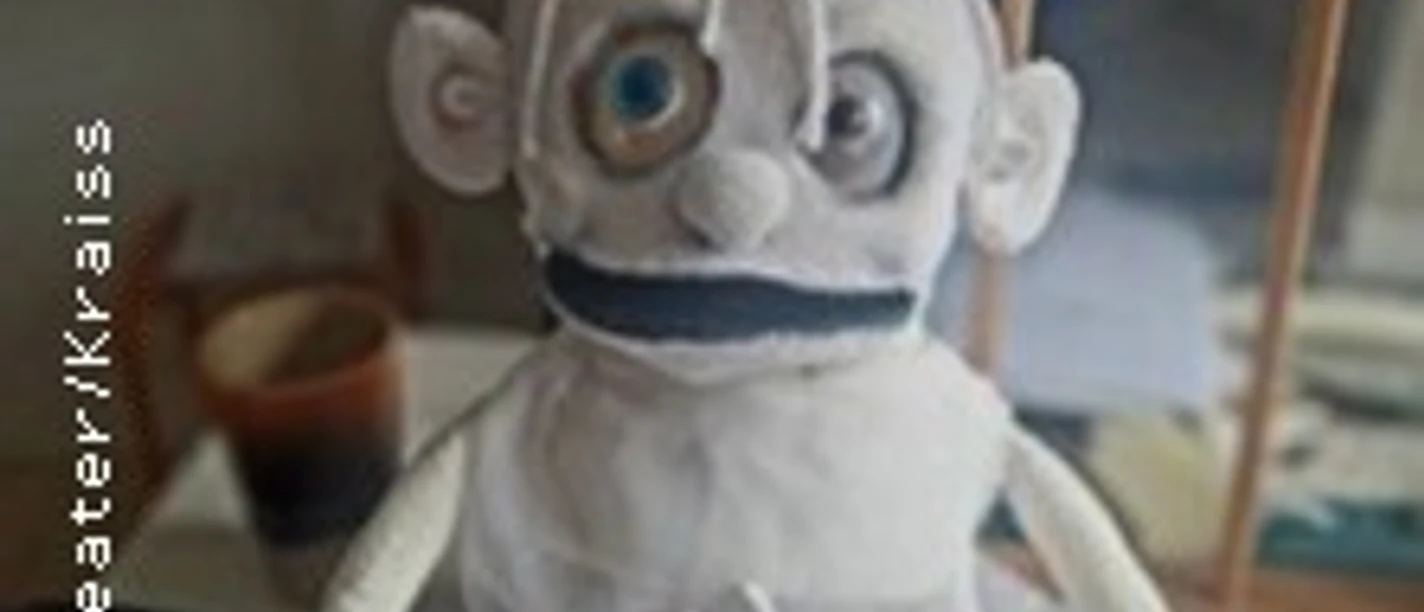 Weiße Stoffpuppe mit großem Auge und kleiner Figur im Schoß vor unscharfem Hintergrund.