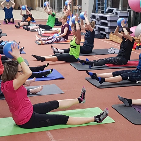 Kräftigung.jpg Aufwärmübungen in der Halle. Die Teilnehmenden sitzen auf Matten und heben Bälle über den Kopf.