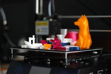 Bunte 3D-Drucke auf einer Plattform: ein Boot, Buchstaben und eine orangefarbene Tierfigur.