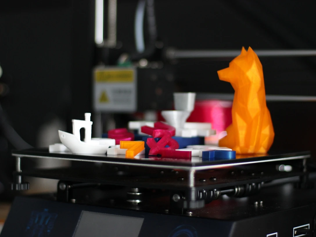 3D-Druck für Kinder Bunte 3D-Drucke auf einer Plattform: ein Boot, Buchstaben und eine orangefarbene Tierfigur.