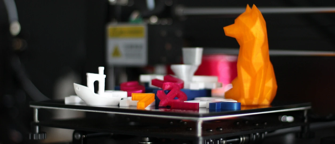 3D-Druck für Kinder Bunte 3D-Drucke auf einer Plattform: ein Boot, Buchstaben und eine orangefarbene Tierfigur.