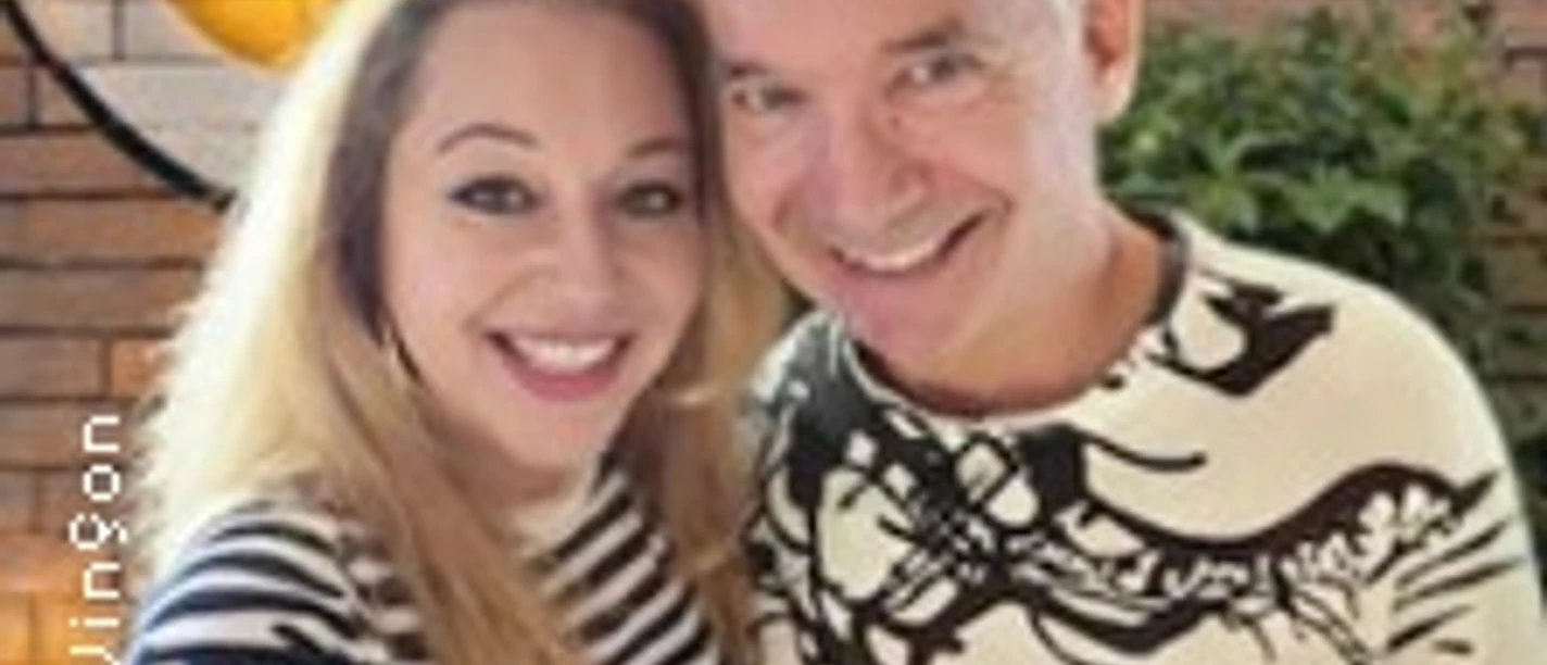 Zwei Personen lächelnd vor Backsteinwand mit großem rundem Wanddekor