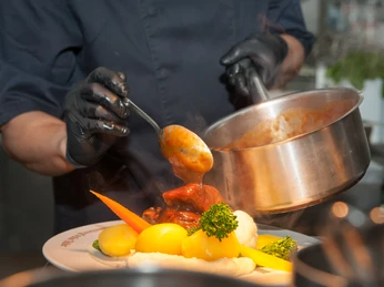 Lauenburgischer Teller 2024 - Kutscherscheune A person pours warm sauce over cooked vegetables and potatoes on a plate.