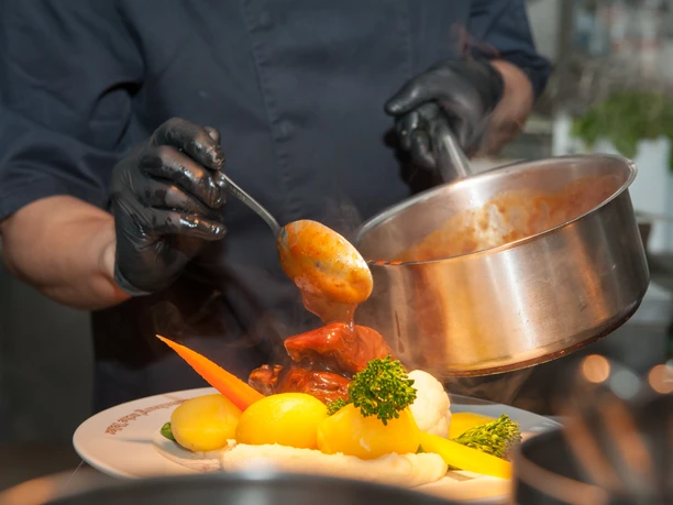 Lauenburgischer Teller 2024 - Kutscherscheune Eine Person gießt warme Soße über gekochtes Gemüse und Kartoffeln auf einem Teller