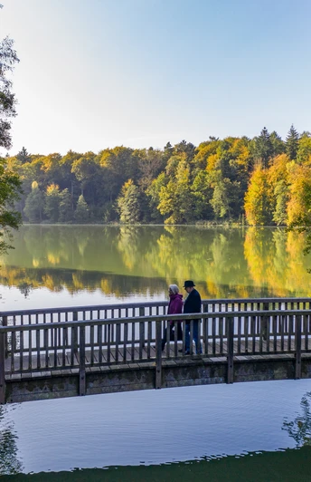 Wandern am Schmalsee Zwei Menschen laufen auf einer Brücke über den Schmalsee bei Mölln. Die umgebenden bäume strahlen in bunten Herbstfarben.
