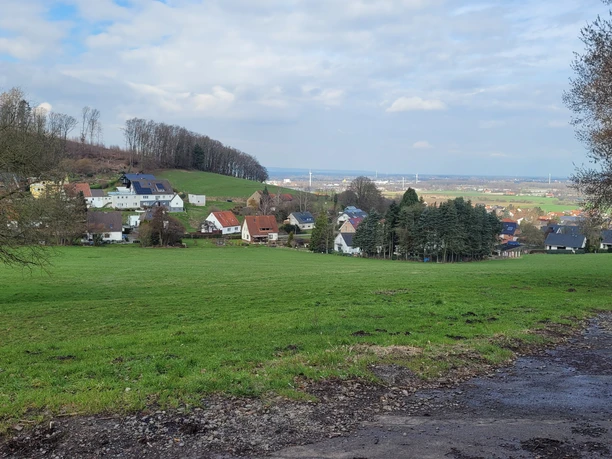 Blick über grüne Wiese auf Häuser am Hang und weite Landschaft unter wolkigem Himmel