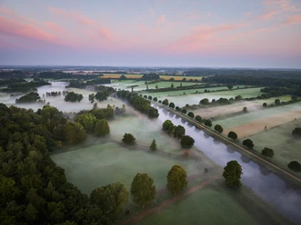 Luftaufnahme vom Elbe-Lübeck-Kanal bei Sonnenaufgang. Nebel liegt über den Feldern und der Himmel ist rosa gefärbt.
