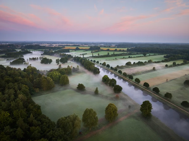 Luftaufnahme vom Elbe-Lübeck-Kanal bei Sonnenaufgang. Nebel liegt über den Feldern und der Himmel ist rosa gefärbt.
