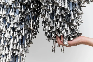 Marie Watt, Thirteen Moons, 2025 Hand hält dicht gebündelte silberne Metallkegel, die an Stoffbändern herabhängen.