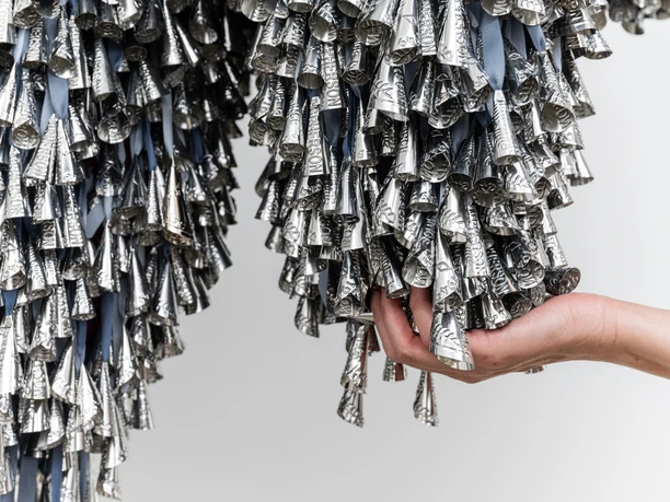 Marie Watt, Thirteen Moons, 2025 Hand hält dicht gebündelte silberne Metallkegel, die an Stoffbändern herabhängen.