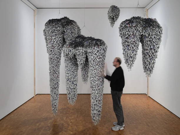 Installation view HERE AND NOW at the Museum Ludwig. De/Collecting Memories Museum Ludwig, Cologne 2026 Hanging sculptural forms made up of many small elements with a blurred person in the room.