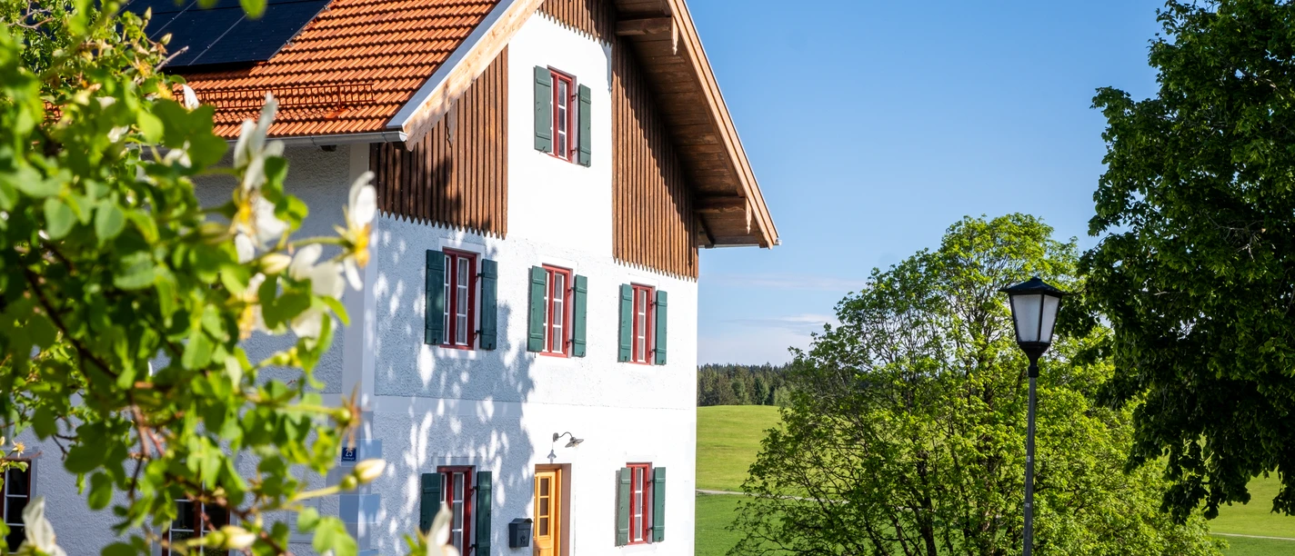 Pilgerhaus Wildsteig Weißes Landhaus mit rotem Dach vor grünen Wiesen und Bäumen an einem sonnigen Tag