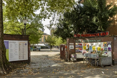 Alte Feuerwache Eingang zur Alten Feuerwache Köln mit Plakatwand, Bäumen und gepflastertem Hofbereich.