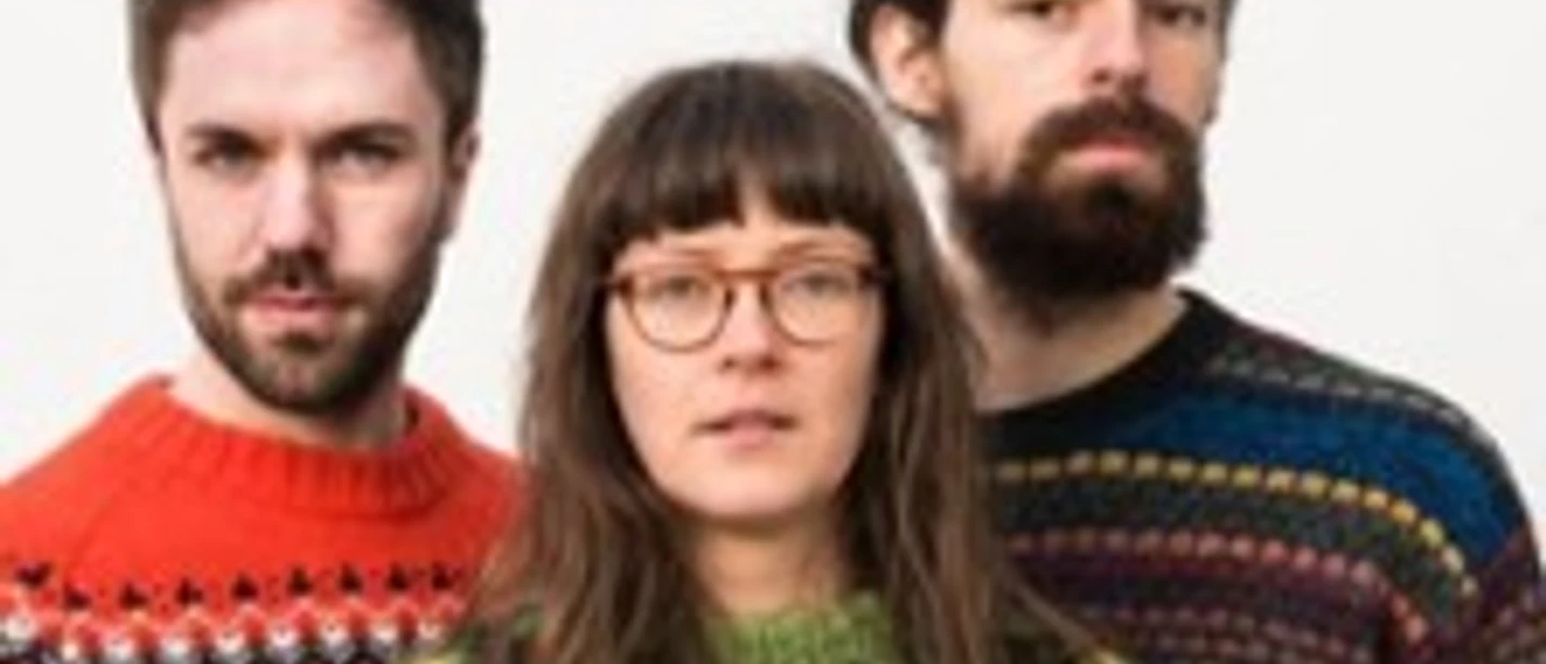 Drei Erwachsene in bunten Strickpullovern stehen eng beieinander vor hellem Hintergrund