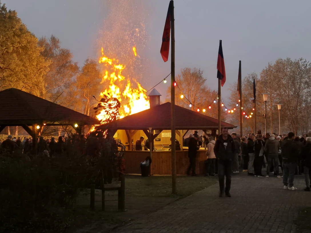 Osterfeuer.jpg Großes Osterfeuer zwischen Holzhütten, umringt von vielen Besuchern in der Abenddämmerung.