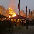 Osterfeuer.jpg Großes Osterfeuer zwischen Holzhütten, umringt von vielen Besuchern in der Abenddämmerung.