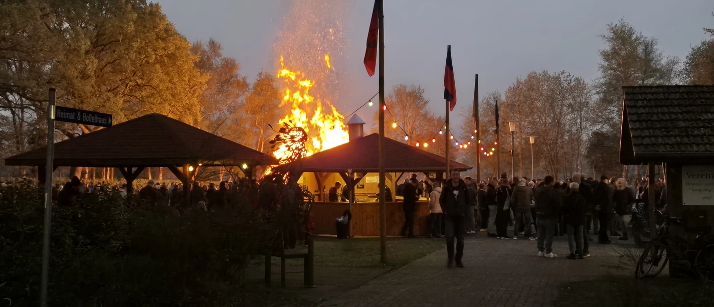 Osterfeuer.jpg Großes Osterfeuer zwischen Holzhütten, umringt von vielen Besuchern in der Abenddämmerung.