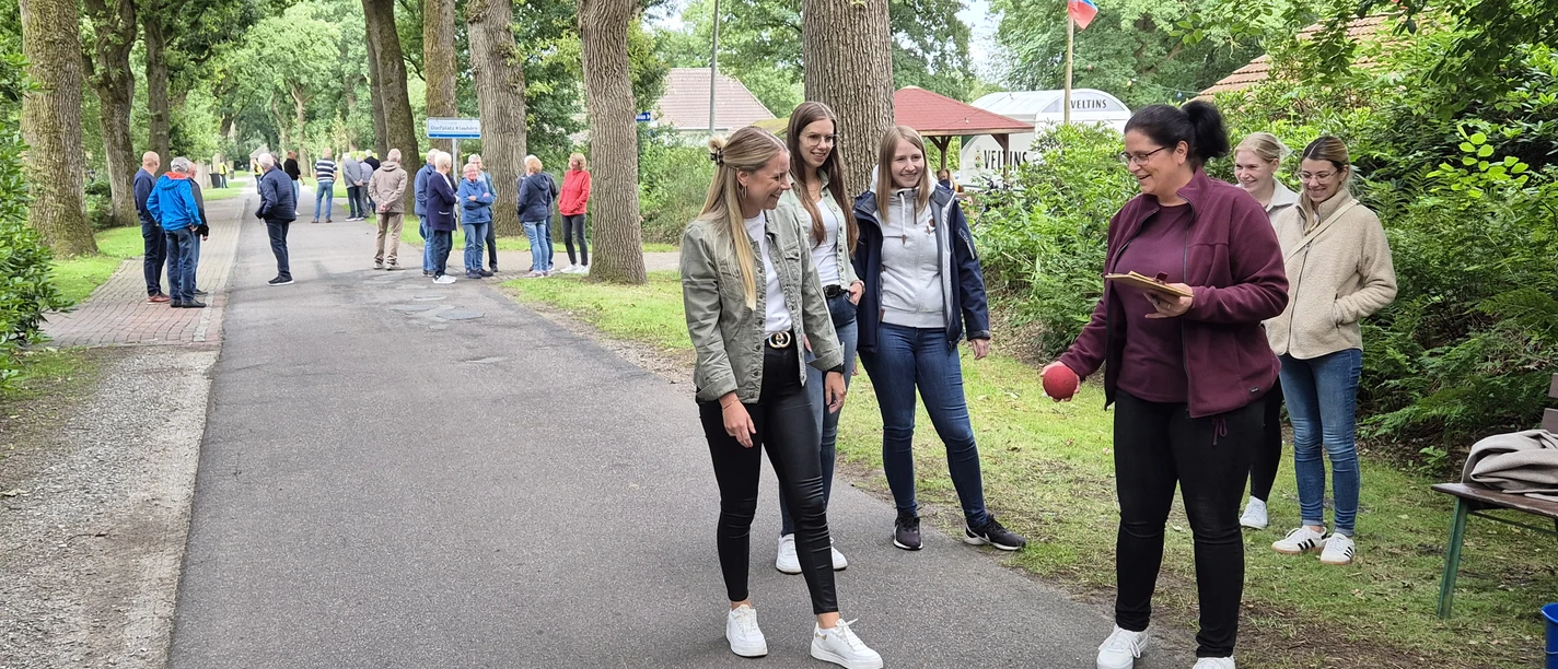 Boßelwochen.jpg Eine fröhliche Frauengruppe beim Boßeln auf einer von Bäumen gesäumten Straße.