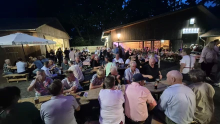 Boßelball.jpg Gesellige Menschenmenge beim Sommerfest auf Bänken zwischen beleuchteten Holzscheunen bei Nacht.