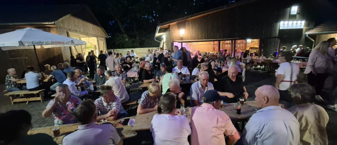 Boßelball.jpg Gesellige Menschenmenge beim Sommerfest auf Bänken zwischen beleuchteten Holzscheunen bei Nacht.