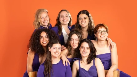 Las Hienas Acht junge Frauen in lila Outfits lächeln vor einem leuchtend orangen Hintergrund.