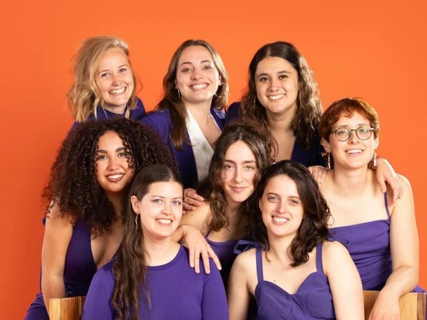 Las Hienas Acht junge Frauen in lila Outfits lächeln vor einem leuchtend orangen Hintergrund.