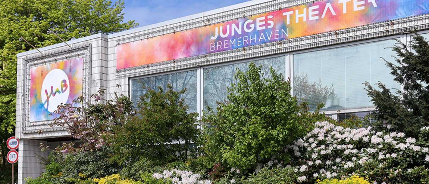 JUB - Junges Theater Bremerhaven_Foto Evelyn Walton_1200x8001692623247.jpg Fassade des Jungen Theaters Bremerhaven mit buntem Banner und blühenden Büschen davor.
